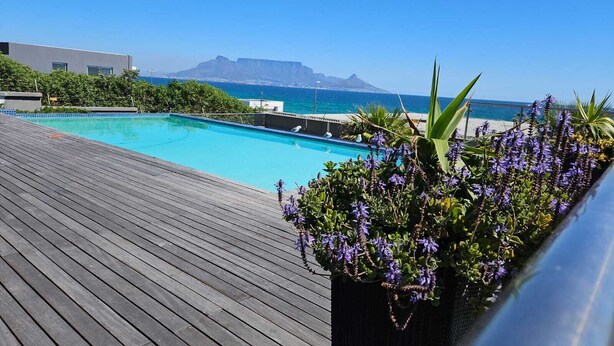 Ein Pool mit Holzterrasse, umgeben von Grün und mit Blick auf einen Strand mit Bergen im Hintergrund.