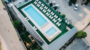 Vista aérea de un hotel con una amplia piscina rectangular, múltiples sillas de descanso y un área de estar cubierta.