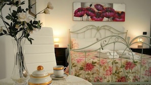 A bedroom with a floral bedspread, a white bedside table with a vase of flowers, and a painting of red flowers on the wall. - B&B Main Street (Reggio Calabria)