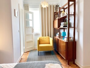 Um corredor com uma poltrona amarela, uma estante de madeira e uma porta que dá para um quarto claro.