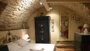 A bedroom with a stone wall, a bed with a white bedspread, a wooden dresser, and a small table with a lamp. - Charming accommodation in a large 16th century building with swimming pool and garden (Vesseaux)