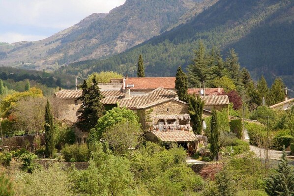 A traditional stone building with terracotta roofs, surrounded by lush greenery and mountains in the background.