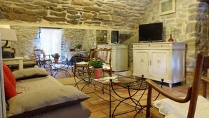 A cozy living room with stone walls, a sofa, a TV, and a dining area with chairs and a table. - Charming accommodation in a large 16th century building with swimming pool and garden (Vesseaux)