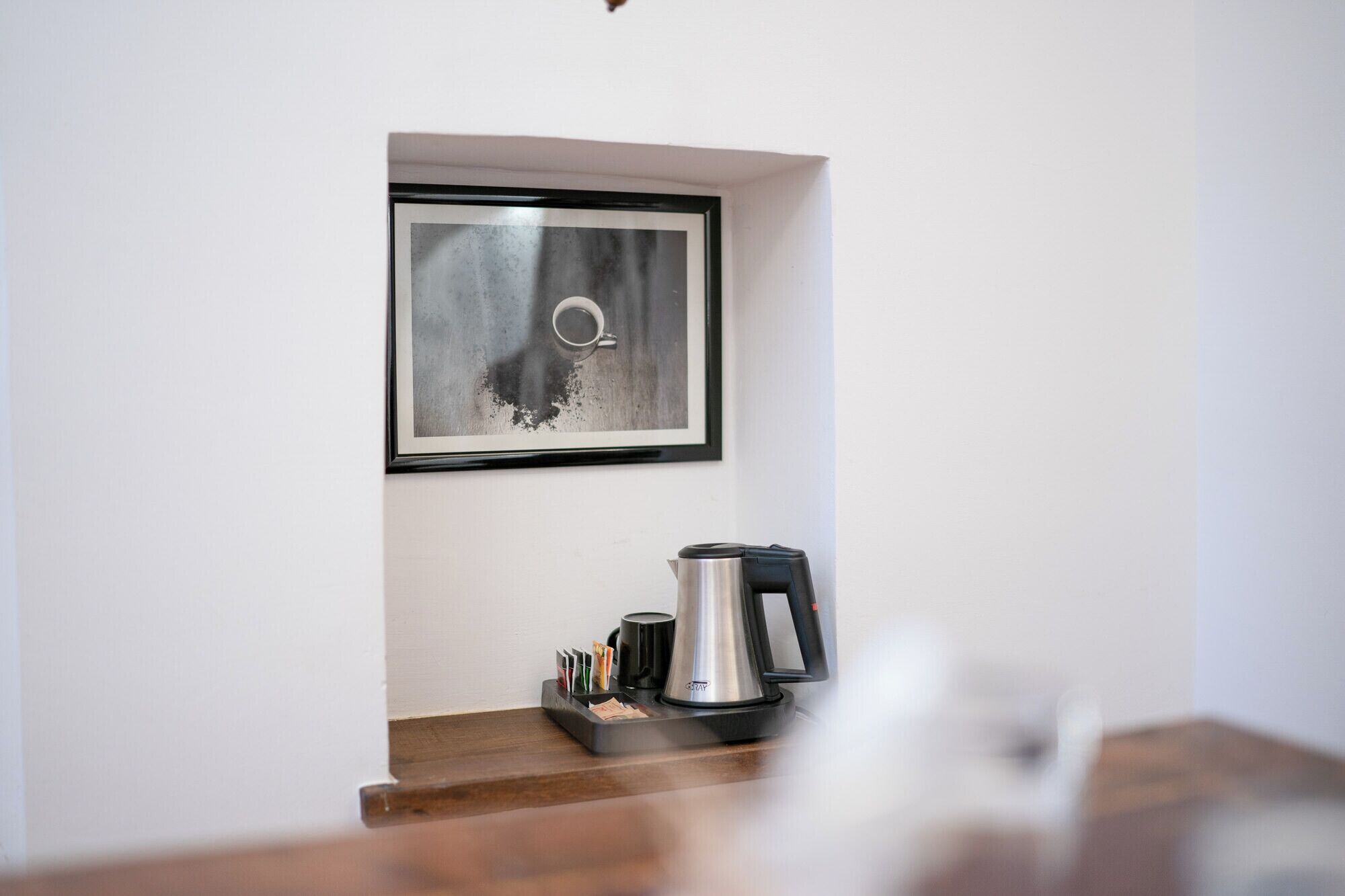 A framed abstract painting on a white wall, a stainless steel coffee maker on a wooden surface, and a black mug.