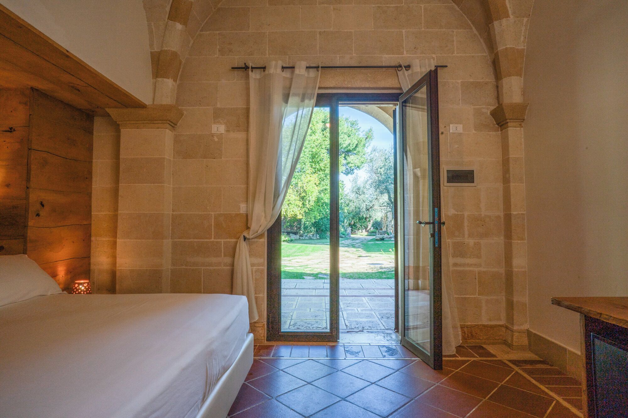 A bedroom with a bed, a window with curtains, and a view of a garden through an open door.