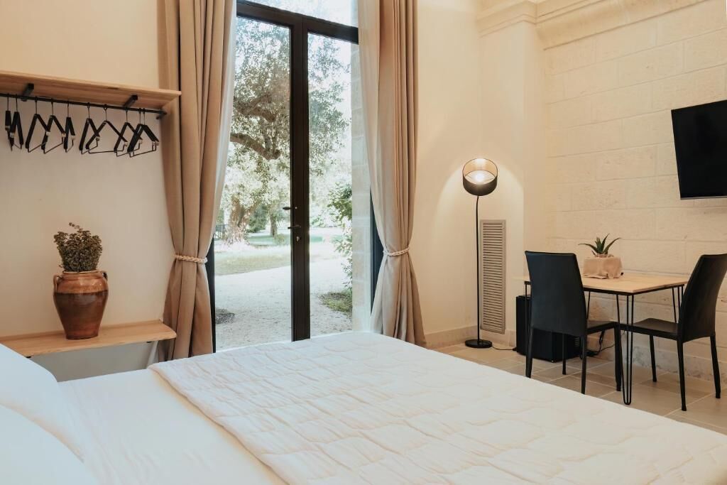 Habitación de hotel con cama, escritorio con sillas, televisión, ventana con cortinas y una planta en maceta.