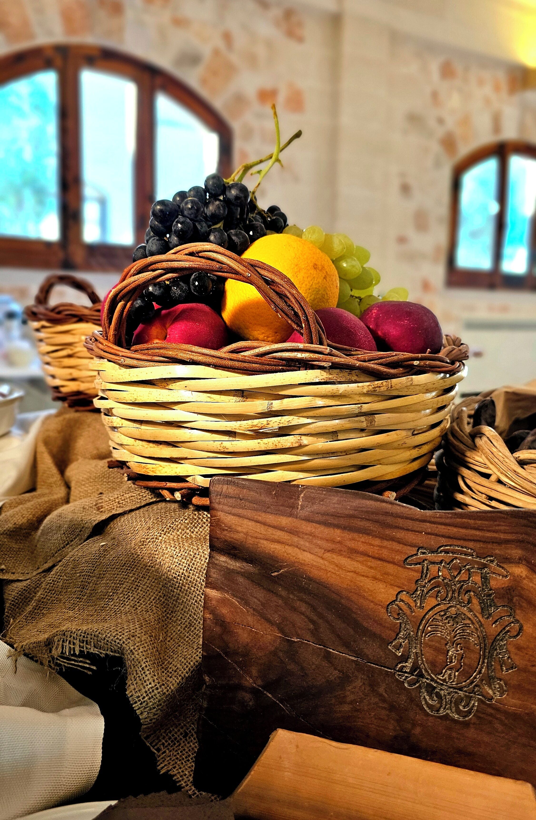 Een rieten mand vol fruit op een tafel met een linnen doek en een houten kist met een versierlijk embleem.