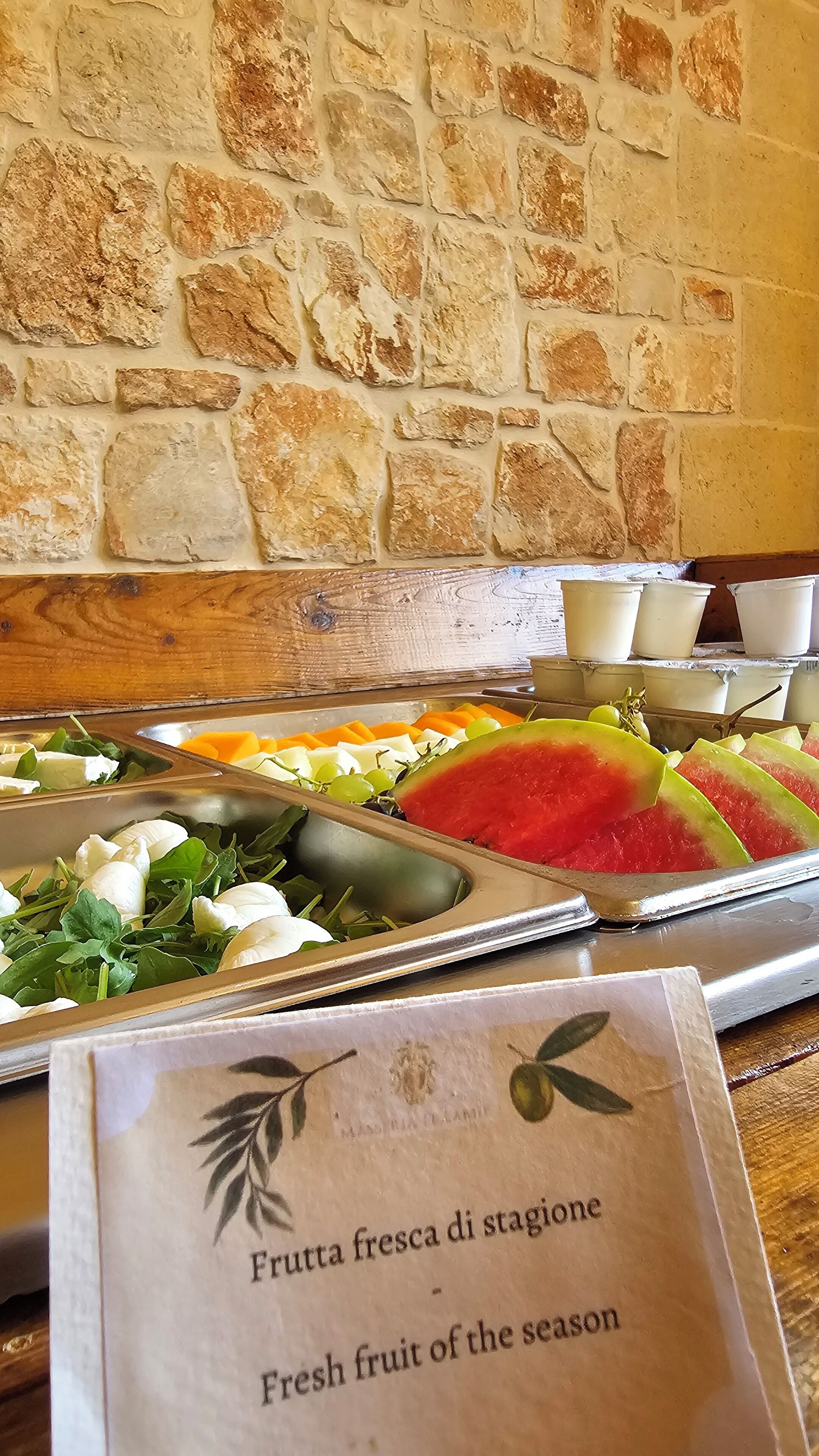 A buffet with various fresh fruits and cheeses, a stone wall background, and a menu sign.