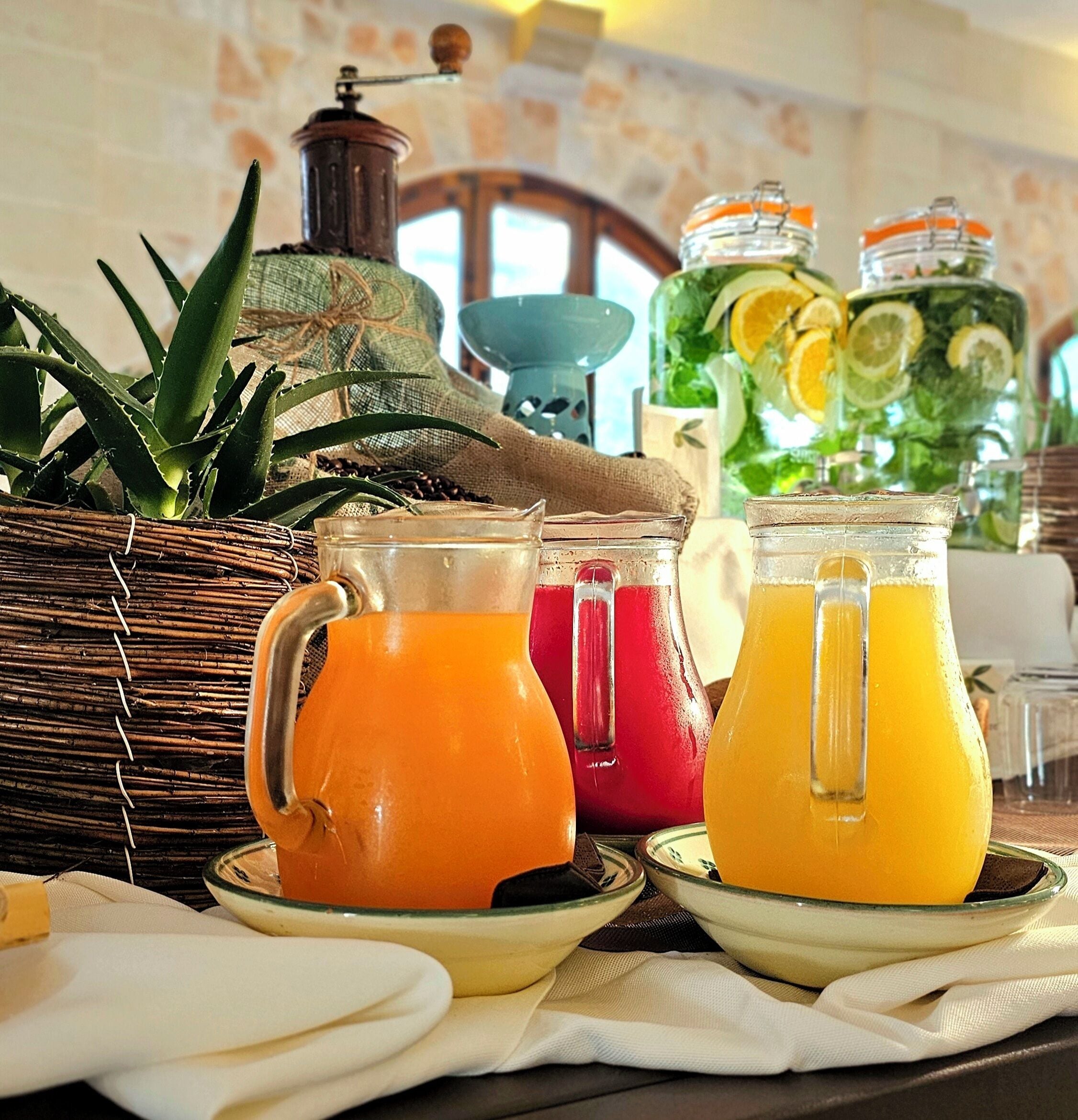 A breakfast setup with three pitchers of orange juice, a wicker basket with green leaves, and a vintage coffee grinder.