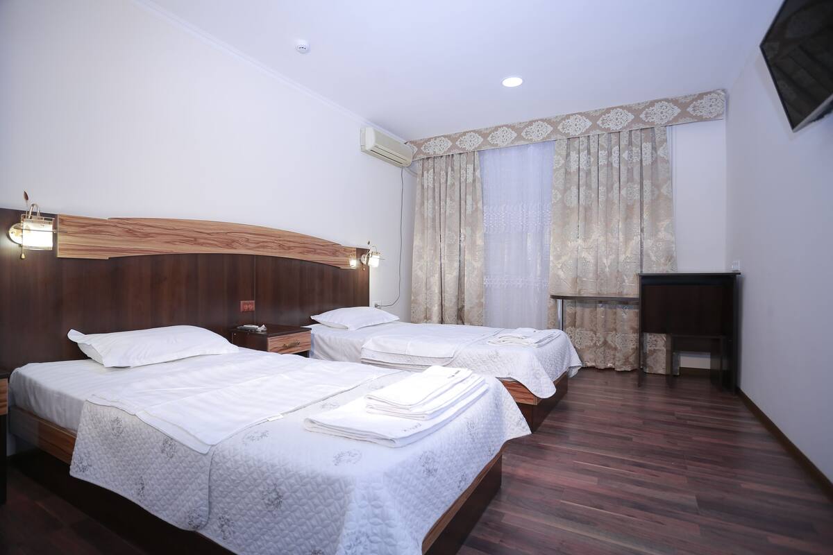A hotel room with two beds, wooden headboards, white bedding, a small bedside table, a nightstand, a desk, and a large window with curtains.