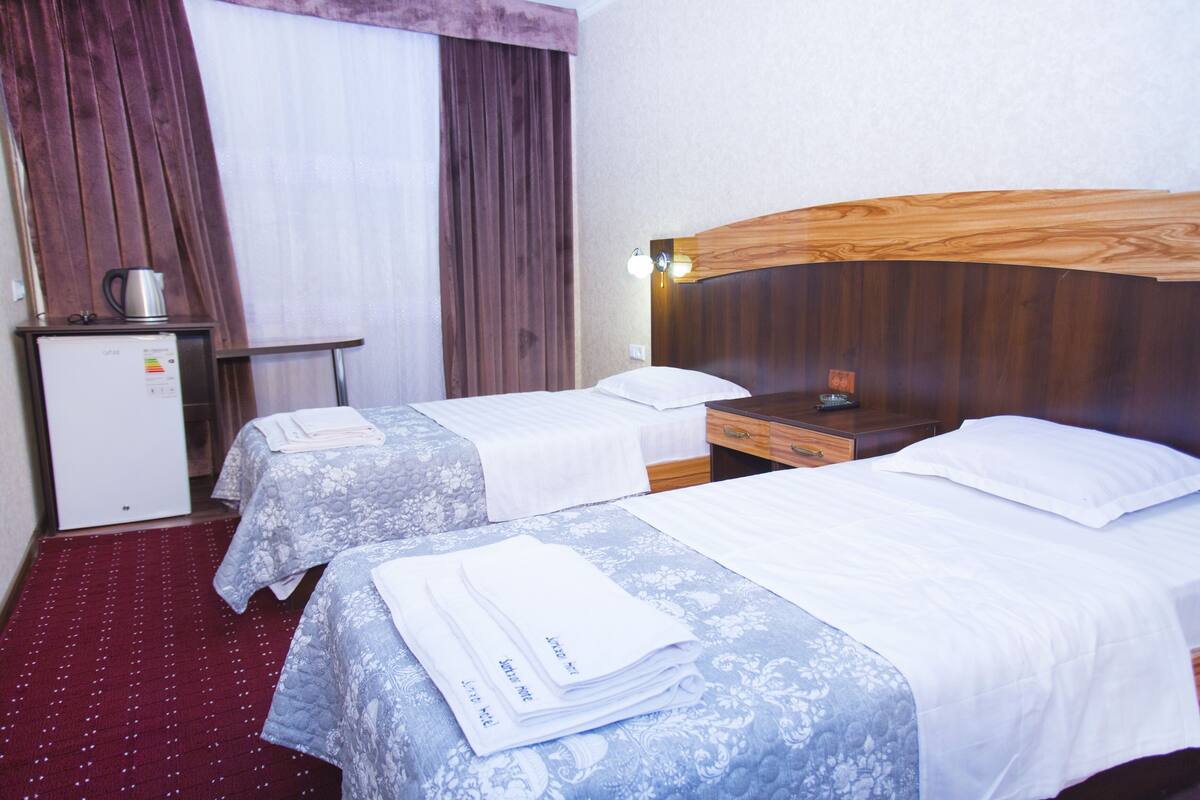 A hotel room with two beds, a small desk with a kettle, and a refrigerator.