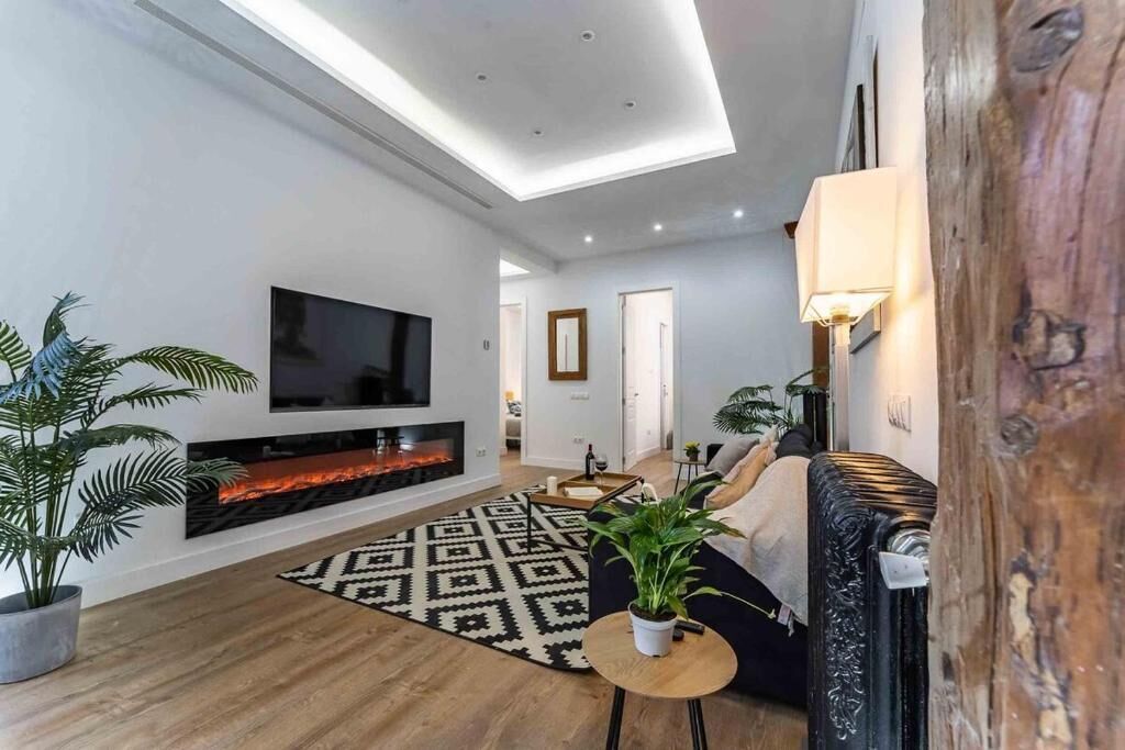 A modern living room with a flat-screen TV, a fireplace, a patterned rug, and a large plant.