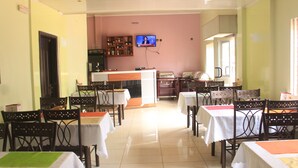 A dining area with tables and chairs, a television mounted on the wall, and a kitchen in the background. - Hotel Exelle-Xior (Yaoundé)