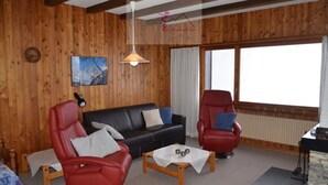 A wooden-paneled room with a black sofa, two red leather chairs, a small table with a white tablecloth, and a large window with curtains. - Nice apartment for rent in les Collons (Thyon-Les Collons)