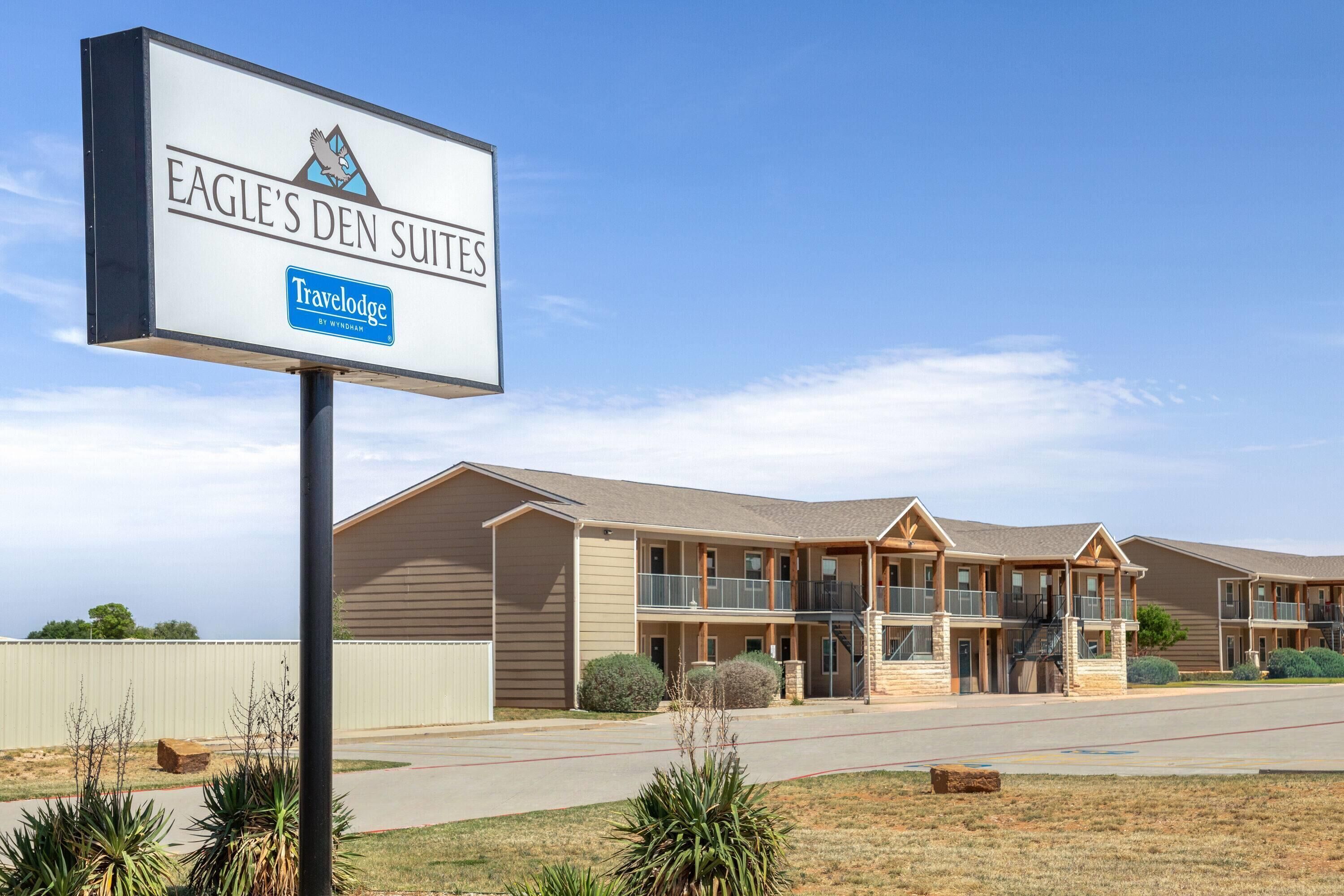 A Travelodge hotel named Eagle's Den Suites with a two-story building, balconies, and a fenced parking area.