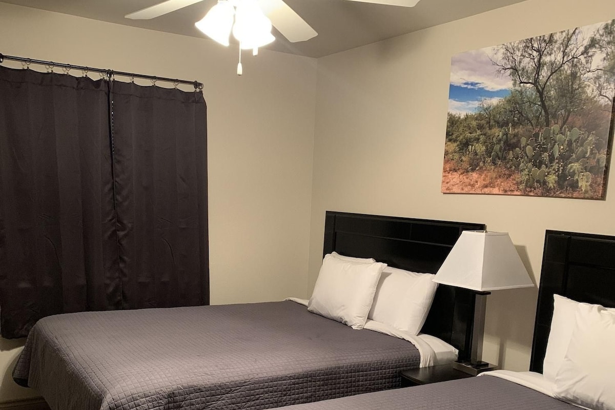 A hotel room with two beds, a ceiling fan, a painting of a desert landscape, and a window with curtains.
