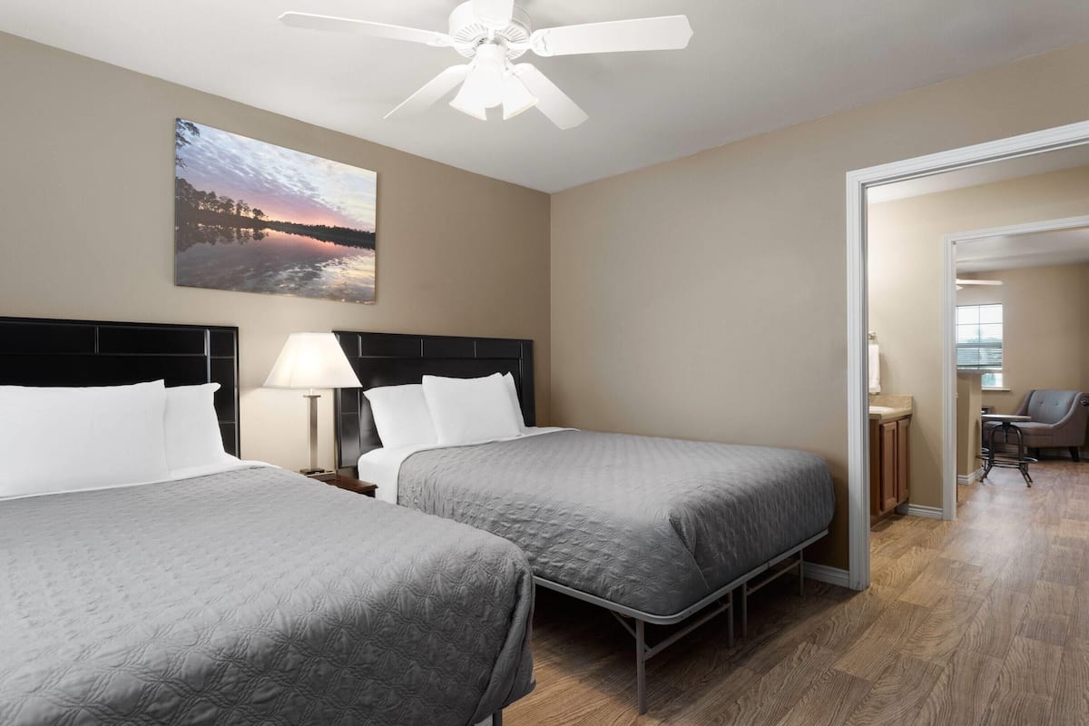 A hotel room with two beds, a ceiling fan, a painting of a sunset, and a view into the adjacent room.
