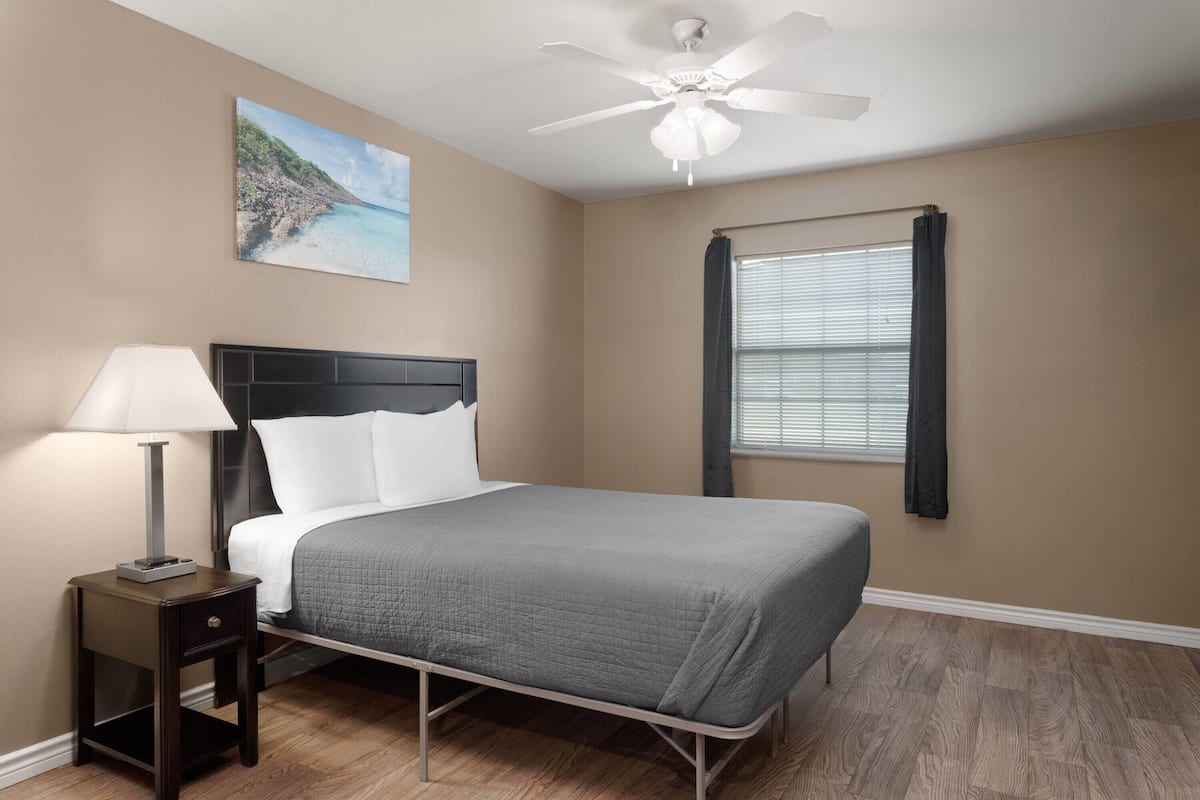 A bedroom with a bed, a nightstand, a ceiling fan, a window with curtains, and a painting on the wall.