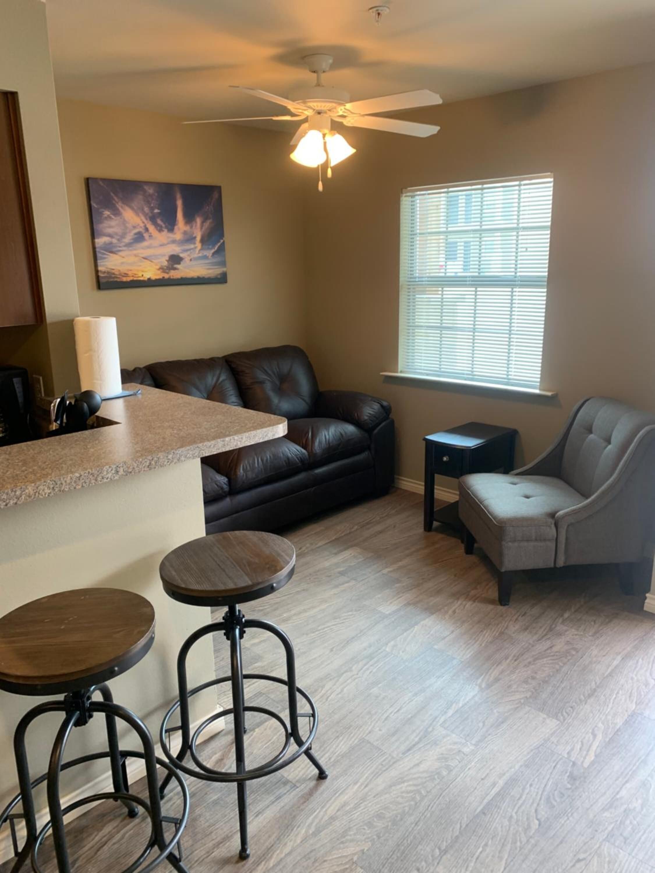 A living room with a black leather sofa, a grey armchair, a small table, and two bar stools. There is a ceiling fan, a window with blinds, and a painting on the wall.