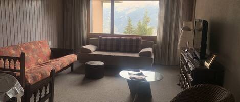 A cozy living room with a sofa, a small round table, a television, and a view of mountains through the window.