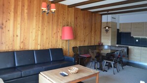 Una habitación con pared de madera, un sofá negro, una mesa de comedor con sillas y una zona de cocina con armarios.