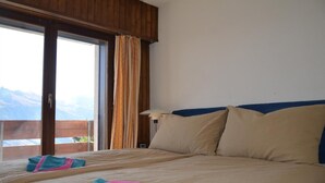 Habitación de hotel con una cama grande, vista a las montañas a través de una puerta corredera y una mesita de noche con lámpara.