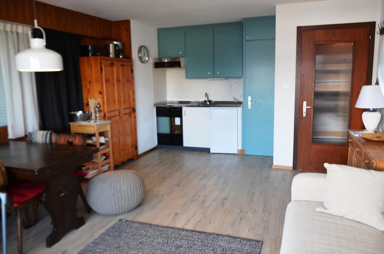 A compact living space with a wooden dining table, a sofa, a kitchen with teal cabinets, and a door with a frosted glass panel.