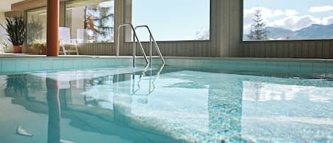 Una piscina interior con agua cristalina, techo de madera y grandes ventanales con vistas a la montaña.