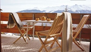 Une terrasse en bois avec une table et des chaises, une théière et des tasses, d’où l’on peut admirer des montagnes enneigées.