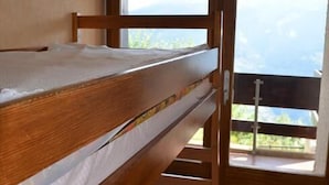 Una cama litera con estructura de madera y ropa de cama blanca, una ventana con vistas a montañas, y un techo de madera con una lámpara circular.