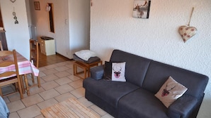 Una sala de estar con un sofá oscuro, una mesa de centro de madera, una mesa de comedor con mantel a cuadros y una mesita auxiliar con un cojín.