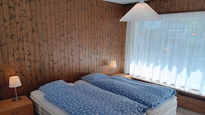 A bedroom with two beds, wooden panel walls, a window with sheer curtains, and a bedside lamp.