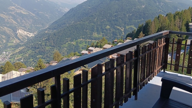 A mountain view from a balcony with a wooden railing, overlooking a valley with houses and a castle-like structure.