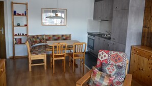 Ett kompakt vardagsrum med ett matbord i trä, två stolar, en soffa med en färgglad kudde, ett inbyggt kök med grå skåp och ett träskåp.