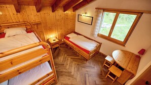 A wooden cabin bedroom with two bunk beds, a single bed, a small table, and a window with a view of greenery.