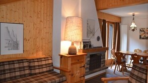 A cozy room with wooden walls, a plaid sofa, a wooden cabinet with a lamp, a dining table with chairs, and a fireplace.