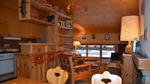 A wooden cabin interior with a kitchen, dining area, and a large window offering a view.