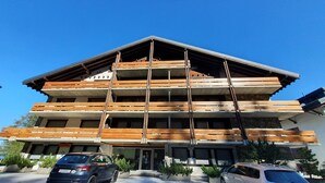 A multi-story building with a sloped roof, wooden balconies, and a parking area with cars.