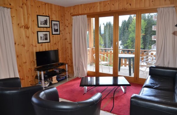 Une pièce aux murs lambrissés, dotée d’un téléviseur, de fauteuils en cuir noir, d’une moquette rouge et d’une porte-fenêtre coulissante donnant sur une terrasse extérieure avec des chaises.