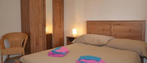 Un dormitorio con un armario de madera, una mesita de noche con lámpara, una cama con dos almohadas y una silla.