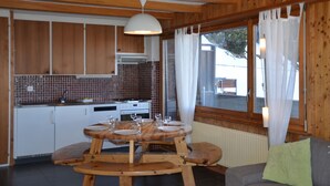 A wooden interior with a dining table set for four, a kitchen with white cabinets, and a view through a window with curtains.