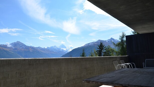 A balcony with a view of snow-capped mountains, a concrete wall, and metal chairs.