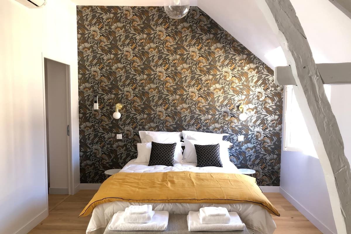 Ein Schlafzimmer mit schrägverdachter Decke, einem Bett mit gelber Tagesdecke, Blumenpapier und einem Nachttisch mit Lampe.