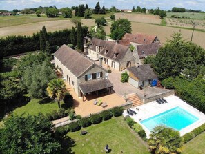 Une grande maison avec une piscine, entourée d’une végétation luxuriante et de champs.