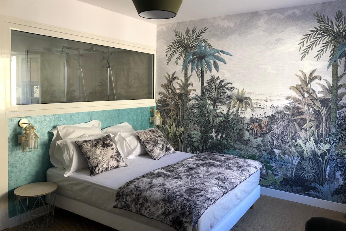 Ein Schlafzimmer mit einem großen Bett, einer Wandmalerei von tropischen Pflanzen, einer modernen Pendelleuchte und einem kleinen runden Tisch.