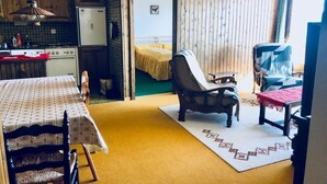 A compact room with a kitchenette, a dining area with a table and chairs, a patterned rug, and a view through an open door to a bed.