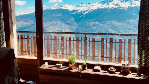 A room with a wooden bench and a small table with a plant, a candle, and a decorative item. A wooden fence is visible outside the window, and a mountain landscape is seen through the window.
