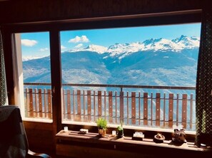 A room with a wooden bench and a small table with a plant, a candle, and a decorative item. A wooden fence is visible outside the window, and a mountain landscape is seen through the window. - On Route 1800, the La Maya building is close to ski lifts and shops. (Thyon-Les Collons)