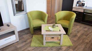 A small, modern room with two green armchairs, a wooden table with a green rug, a small basket, and a kitchen area with appliances. - Ferienwohnung Anke-Apartement 3 (Heinsberg)