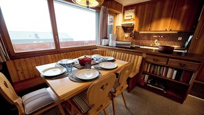 A wooden dining table set with plates, glasses, and a bowl. There are chairs with striped cushions. A bookshelf with books is to the right.